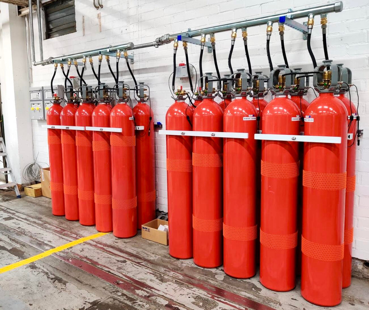 A set of red cylindrical fire extinguisher cylinders mounted on a white brick wall with a metal piping system above them, photographed in an industrial room.