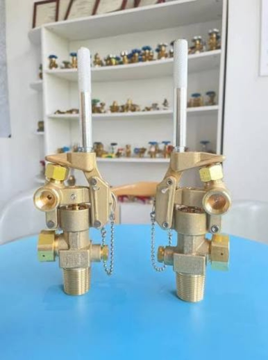 Carbon Dioxide CO2 Fire Suppression System Sleeve Valves. Two brass sprinkler nozzles with white handles and safety chains, displayed on a blue table with a white shelving unit in the background.